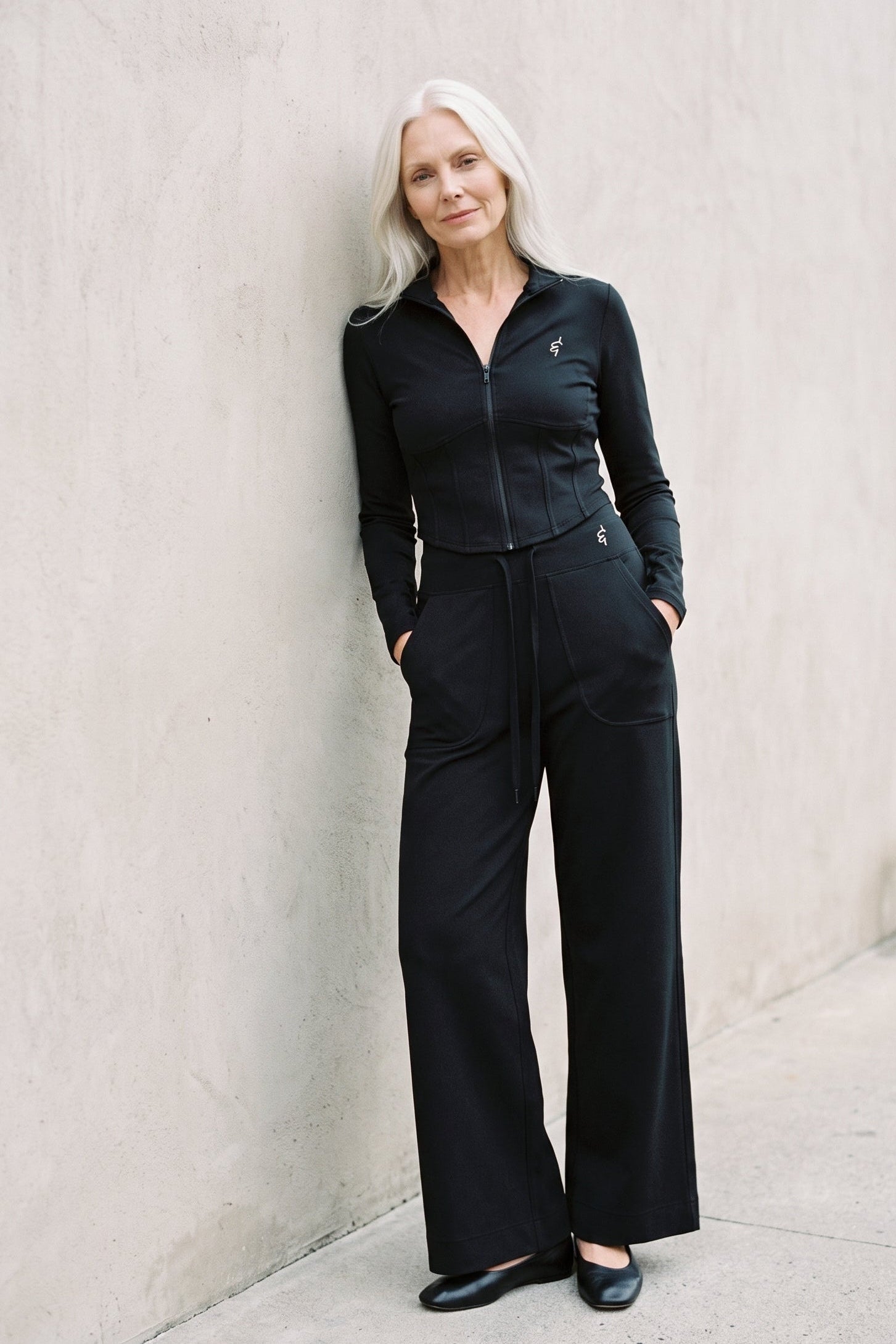 A woman with platinum blonde hair stands against a textured concrete wall wearing a black zip-up long-sleeve top and matching wide-leg trousers.