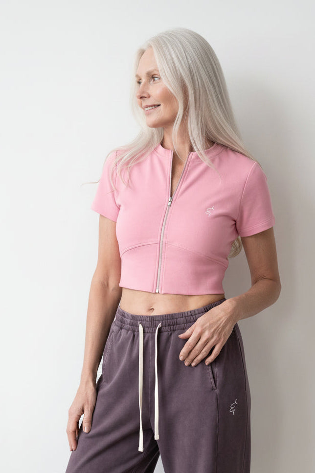 A woman with long white hair stands against a plain light gray wall wearing a pink short-sleeve cropped zip-front top and dark mauve drawstring sweatpants.