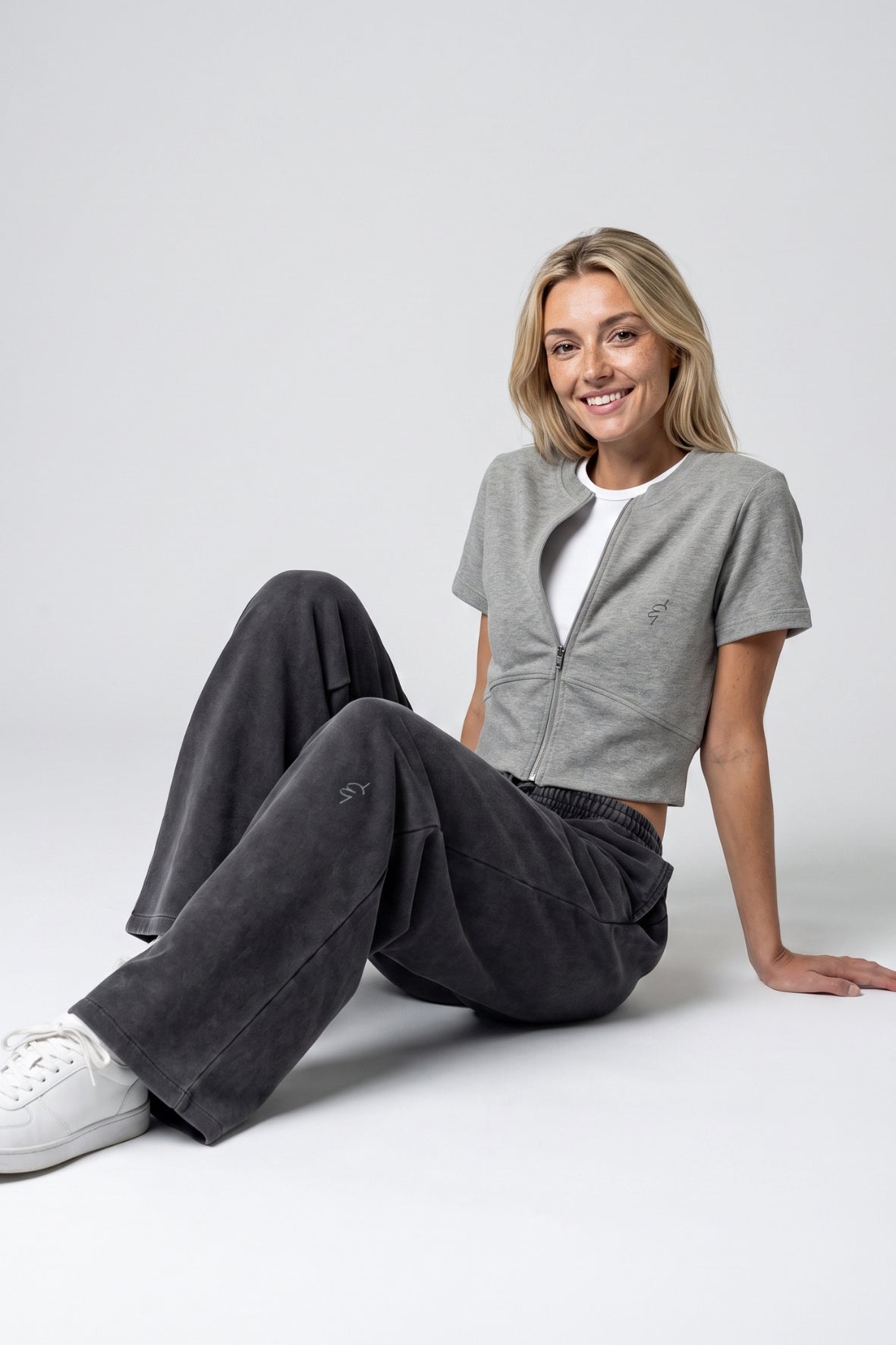 A woman sitting on a light gray studio floor wearing a light gray cropped zip-up cardigan over a white t-shirt, dark gray wide-leg pants, and white sneakers.