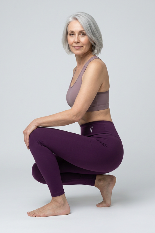 A woman with short gray hair crouches in a yoga pose, wearing a mauve sports bra and dark purple high-waisted leggings against a light gray studio background.