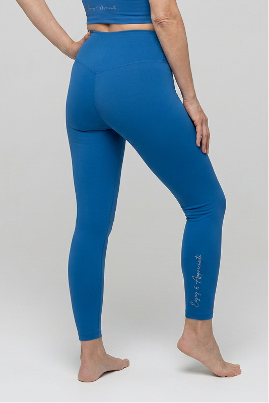 A woman with short gray hair is shown from the back wearing a blue athletic set consisting of a short-sleeve crop top and high-waisted leggings, standing against a light gray studio background.