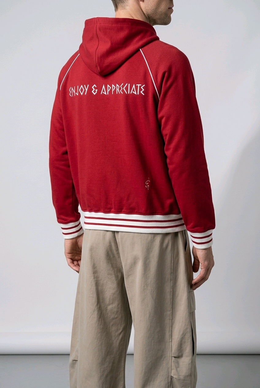 A man shown from the back wearing a red hooded sweatshirt with white text reading 'ENJOY & APPRECIATE' across the upper back, plus white and red striped ribbed cuffs and hem, paired with beige pants against a plain light gray studio background.