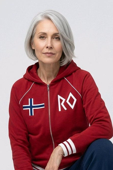 A woman with gray hair crouching in a studio setting, wearing a red zip-up hoodie with a white Norwegian flag and 'RO' graphic on the chest, navy blue pants, and white sneakers.