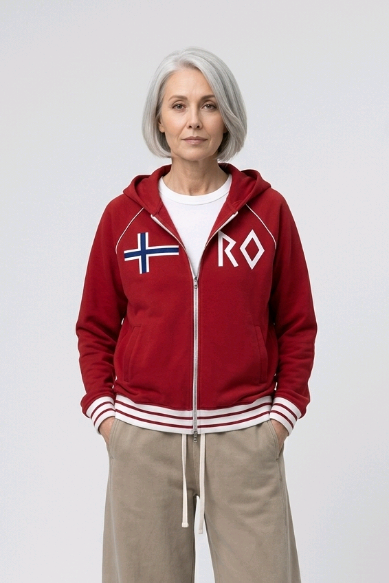 An older woman with gray hair stands against a light gray studio background wearing a red zip-up hoodie with white and blue Nordic flag graphics on the chest, beige wide-leg sweatpants, and white sneakers.