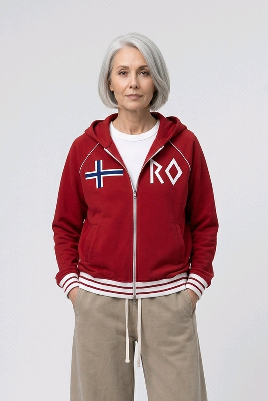 An older woman with gray hair stands against a light gray studio background wearing a red zip-up hoodie with white and blue Nordic flag graphics on the chest, beige wide-leg sweatpants, and white sneakers.