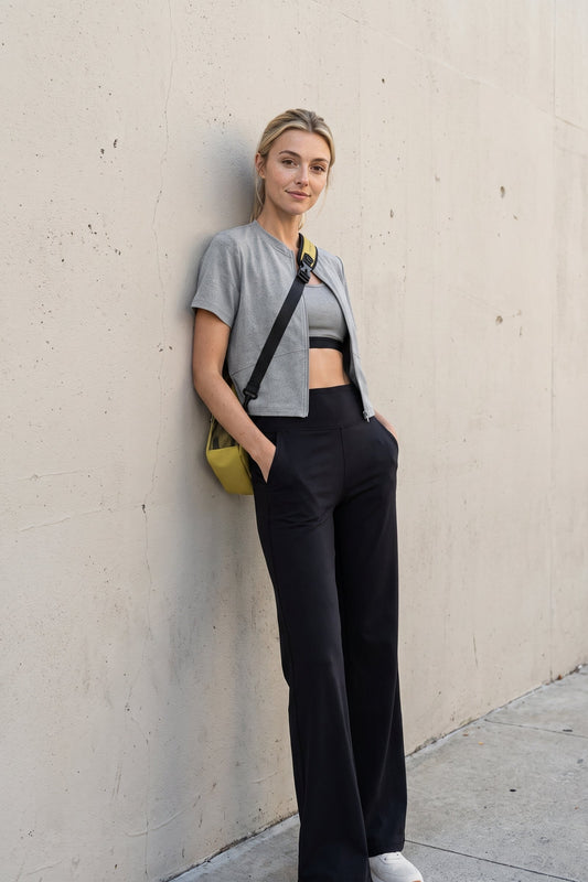 A woman stands against a concrete wall wearing a gray cropped t-shirt, a black sports bra, black wide-leg trousers, and white sneakers, with a small yellow crossbody bag.