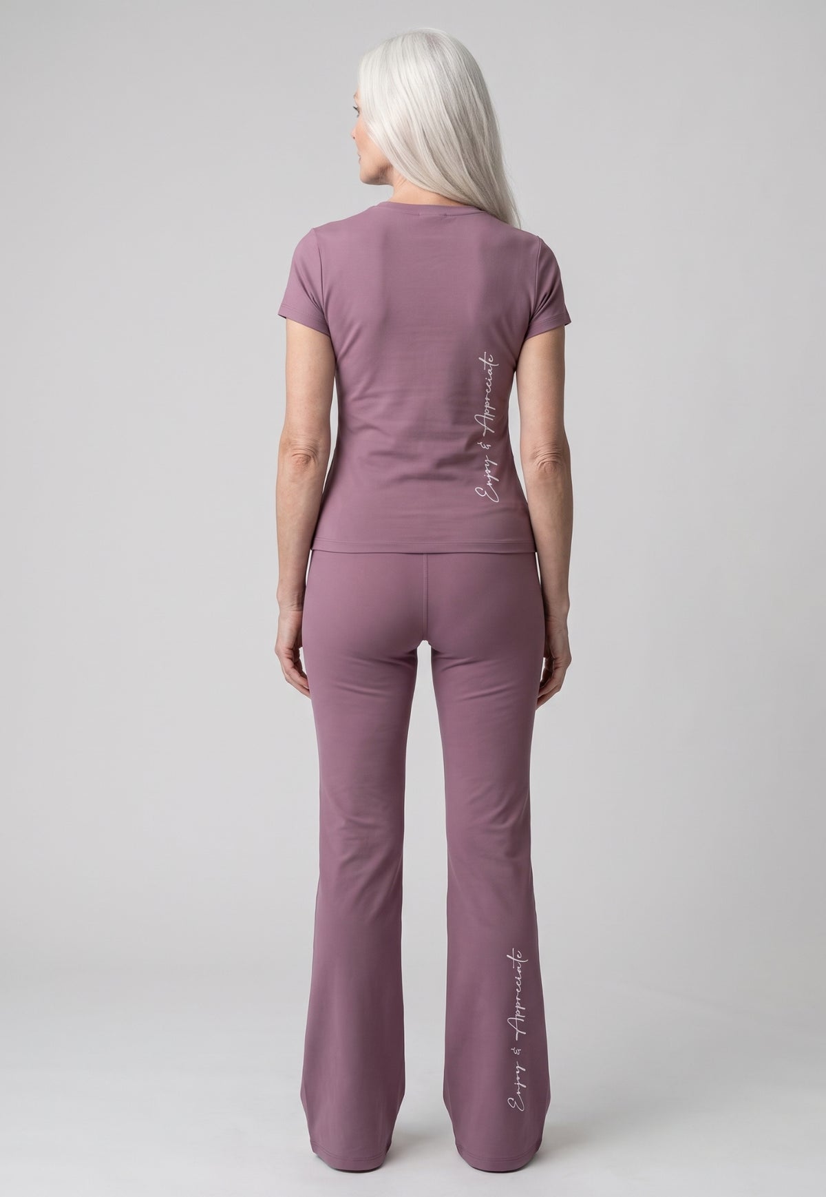 A woman with long platinum blonde hair is shown from the back wearing a matching mauve purple scrub set with a short-sleeve top and flared pants. The set features white script text running vertically down the back and left leg.