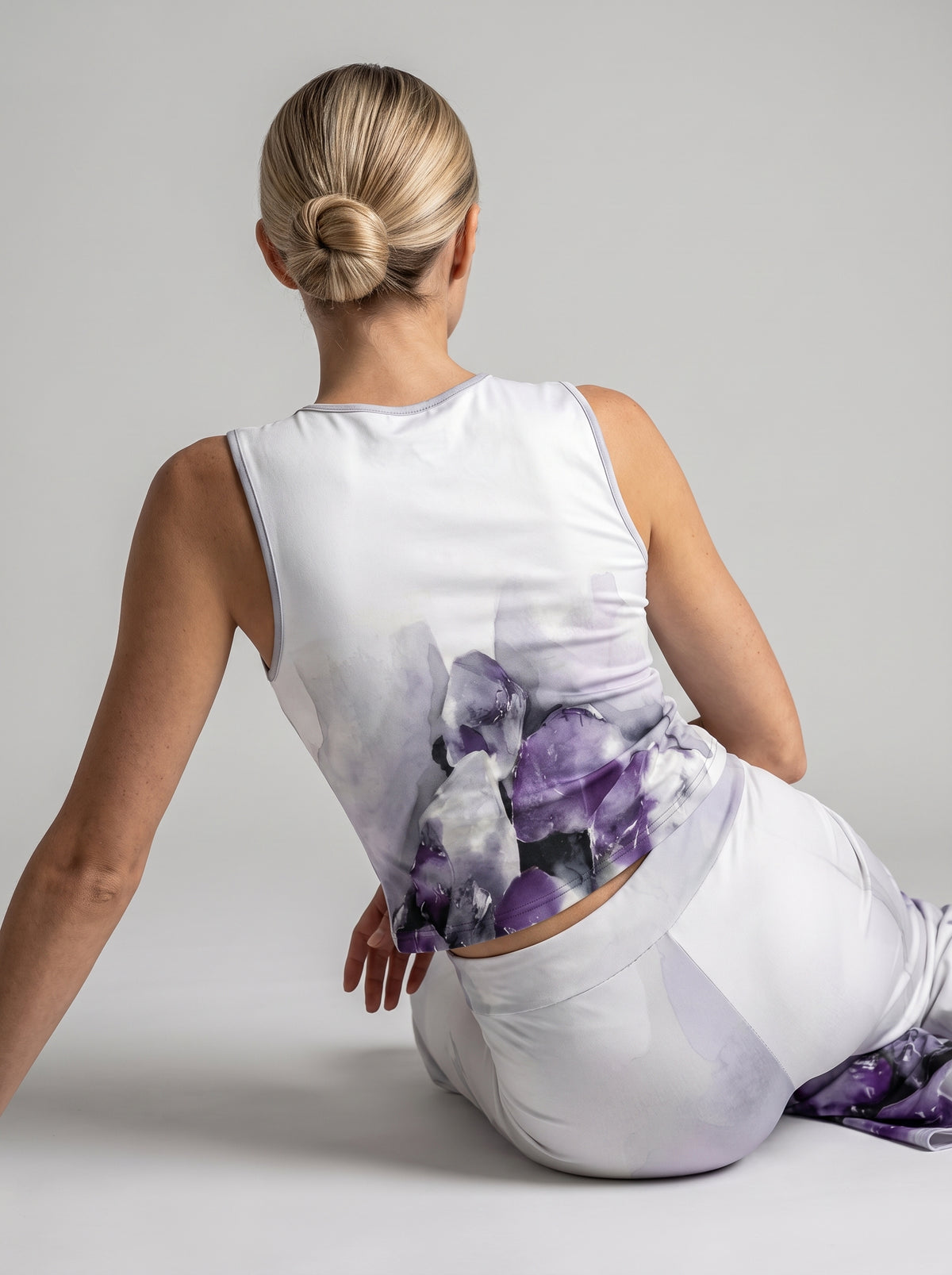 A woman sitting on a light gray studio floor, viewed from behind, wearing a white sleeveless activewear top and matching leggings with a large purple and gray crystal-like abstract print concentrated on the back and lower area.