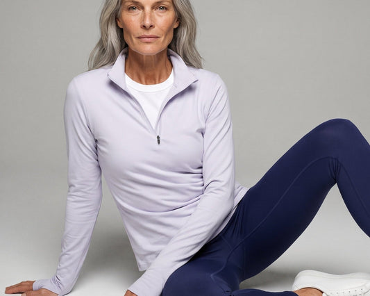 A woman with gray hair sits on a light gray studio floor wearing a light lavender quarter-zip athletic pullover, navy blue leggings, and white sneakers.