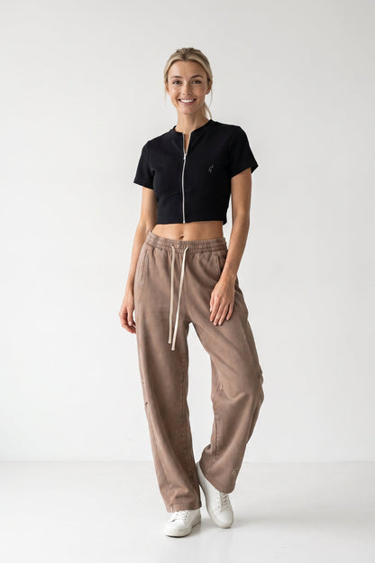 A woman modeling a black cropped zip-up t-shirt and brown wide-leg sweatpants with white sneakers, standing against a plain white studio background.