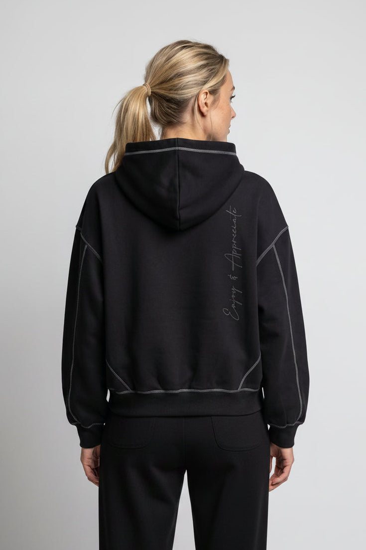 A woman viewed from the back wearing a black cropped hoodie with subtle light gray piping and script text on the back, paired with dark gray pants, against a light gray studio background.