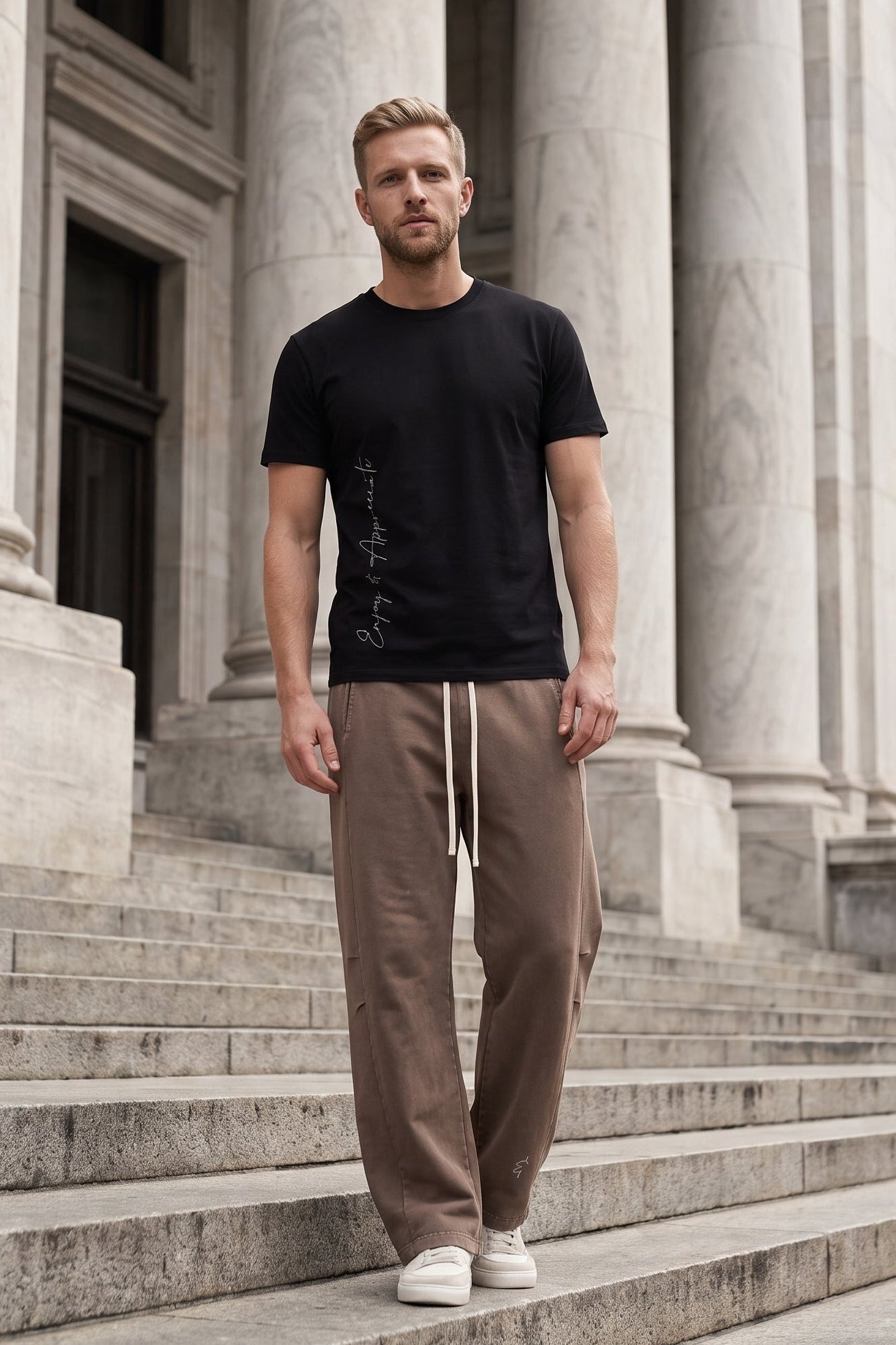 A man stands on stone steps in front of a classical building with large columns, wearing a black short-sleeve t-shirt with a vertical graphic print, brown drawstring sweatpants, and white sneakers.