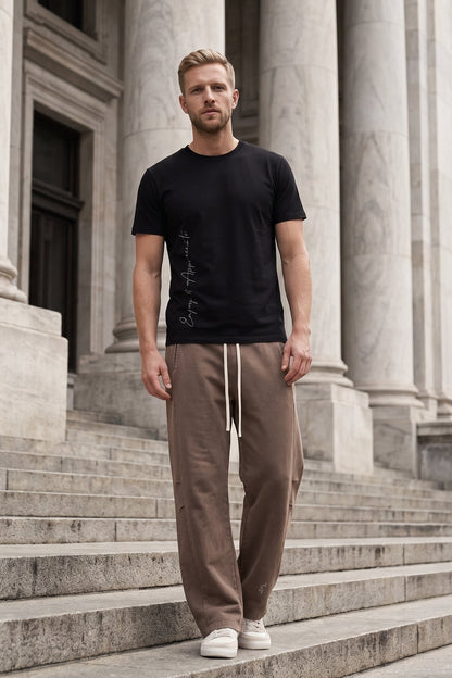 A man stands on stone steps in front of a classical building with large columns, wearing a black short-sleeve t-shirt with a vertical graphic print, brown drawstring sweatpants, and white sneakers.