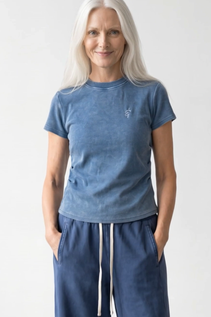 A woman with long white hair wearing a washed slate blue short-sleeve t-shirt and matching blue sweatpants with white drawstrings, standing against a light gray studio background.