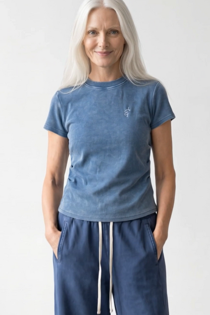 A woman with long white hair wearing a washed slate blue short-sleeve t-shirt and matching blue sweatpants with white drawstrings, standing against a light gray studio background.