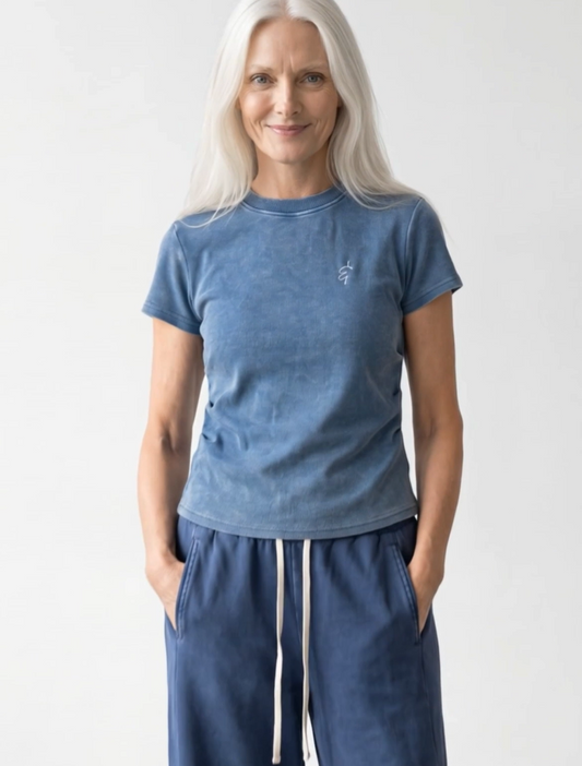 A woman with long white hair wearing a washed slate blue short-sleeve t-shirt and matching blue sweatpants with white drawstrings, standing against a light gray studio background.
