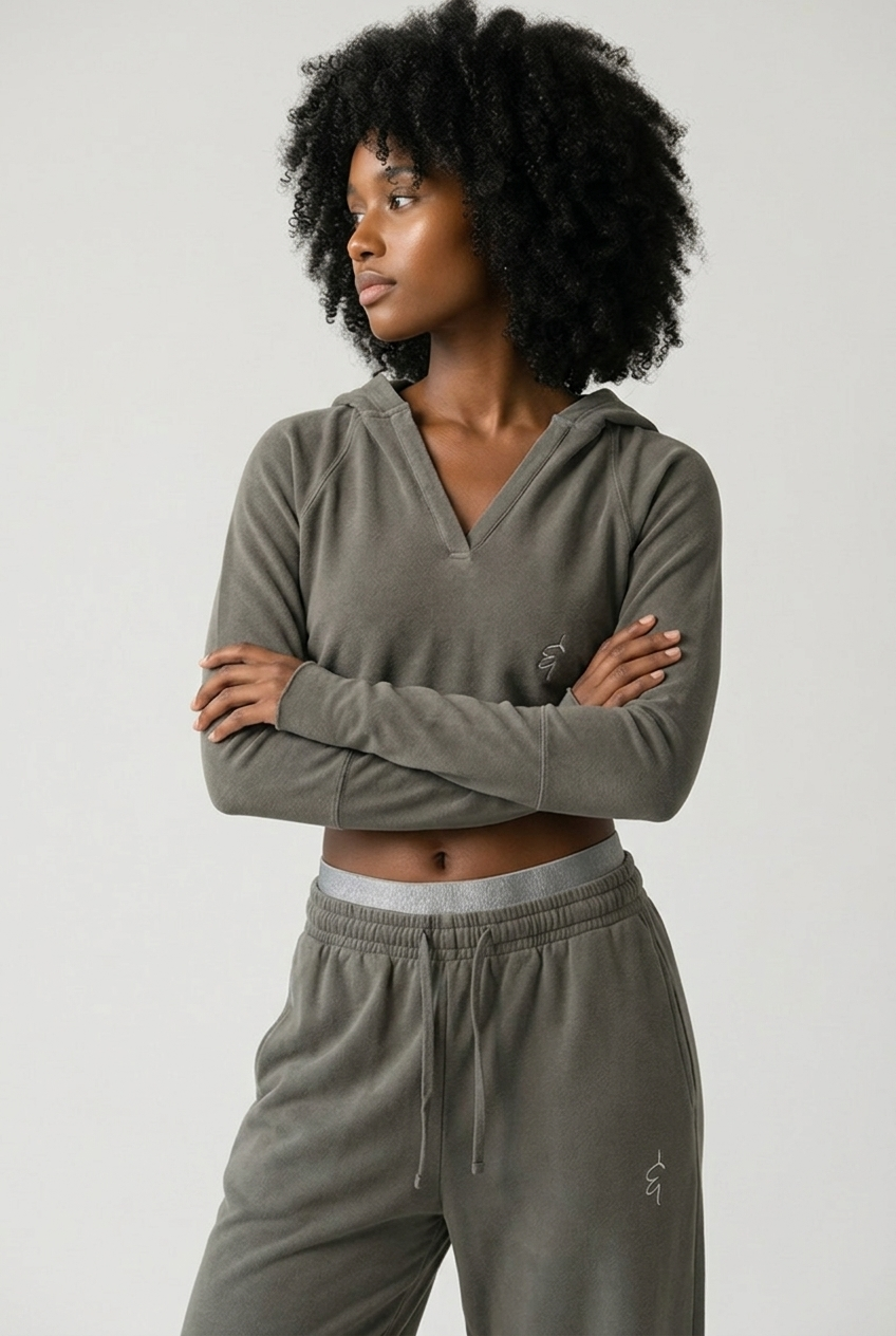 A woman with curly black hair wearing a matching gray-green hooded sweatshirt and jogger sweatpants, standing with her arms crossed against a light gray studio background.