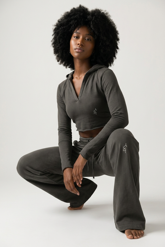 A model with an afro hairstyle crouches in a studio setting, wearing a dark gray long-sleeve crop top and matching sweatpants.