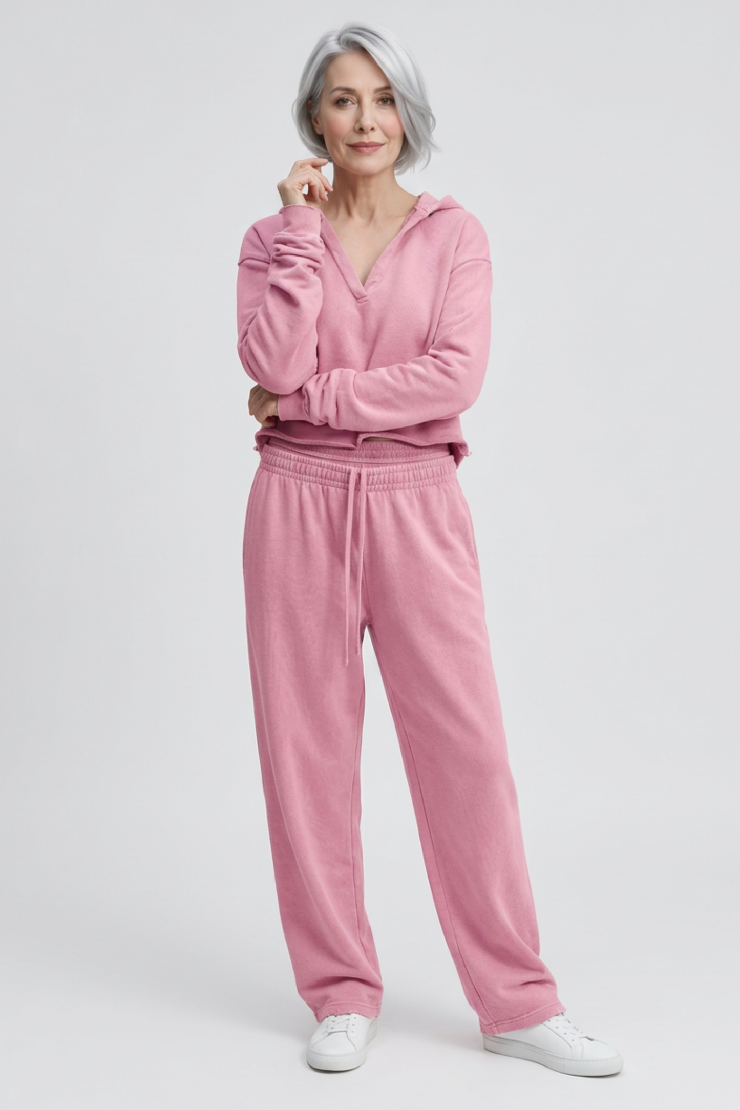 A woman with silver-gray hair wearing a matching pink lounge set consisting of a long-sleeve V-neck hoodie and wide-leg sweatpants, paired with white sneakers, against a light gray studio background.
