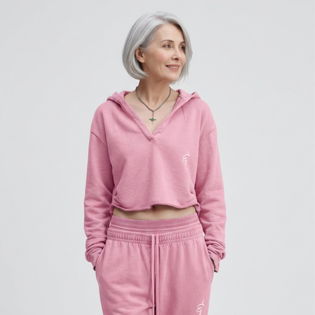 A woman with short gray hair wearing a pink cropped hooded sweatshirt and matching pink sweatpants, photographed against a light gray studio background.