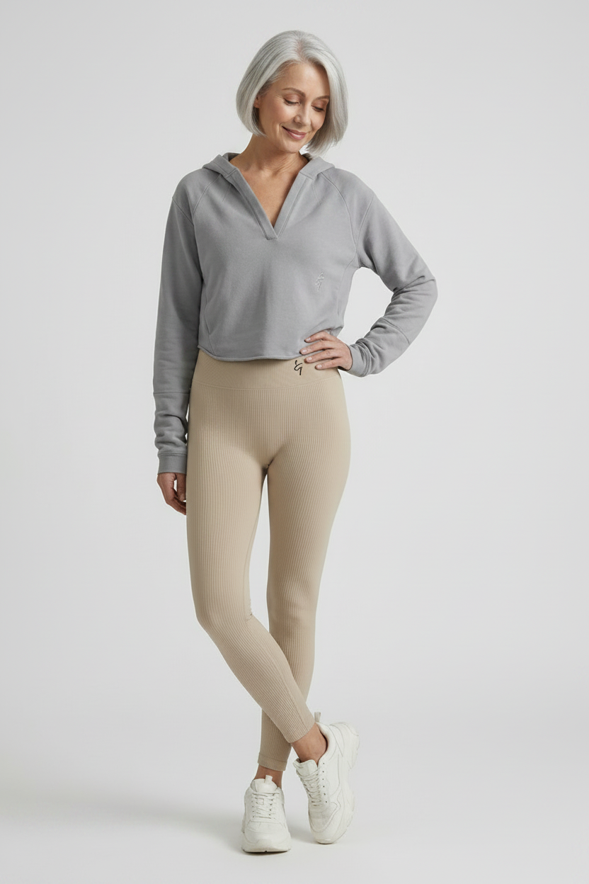 A woman with short gray hair stands in a studio wearing a light gray cropped long-sleeve top, beige leggings, and white sneakers against a light gray background.