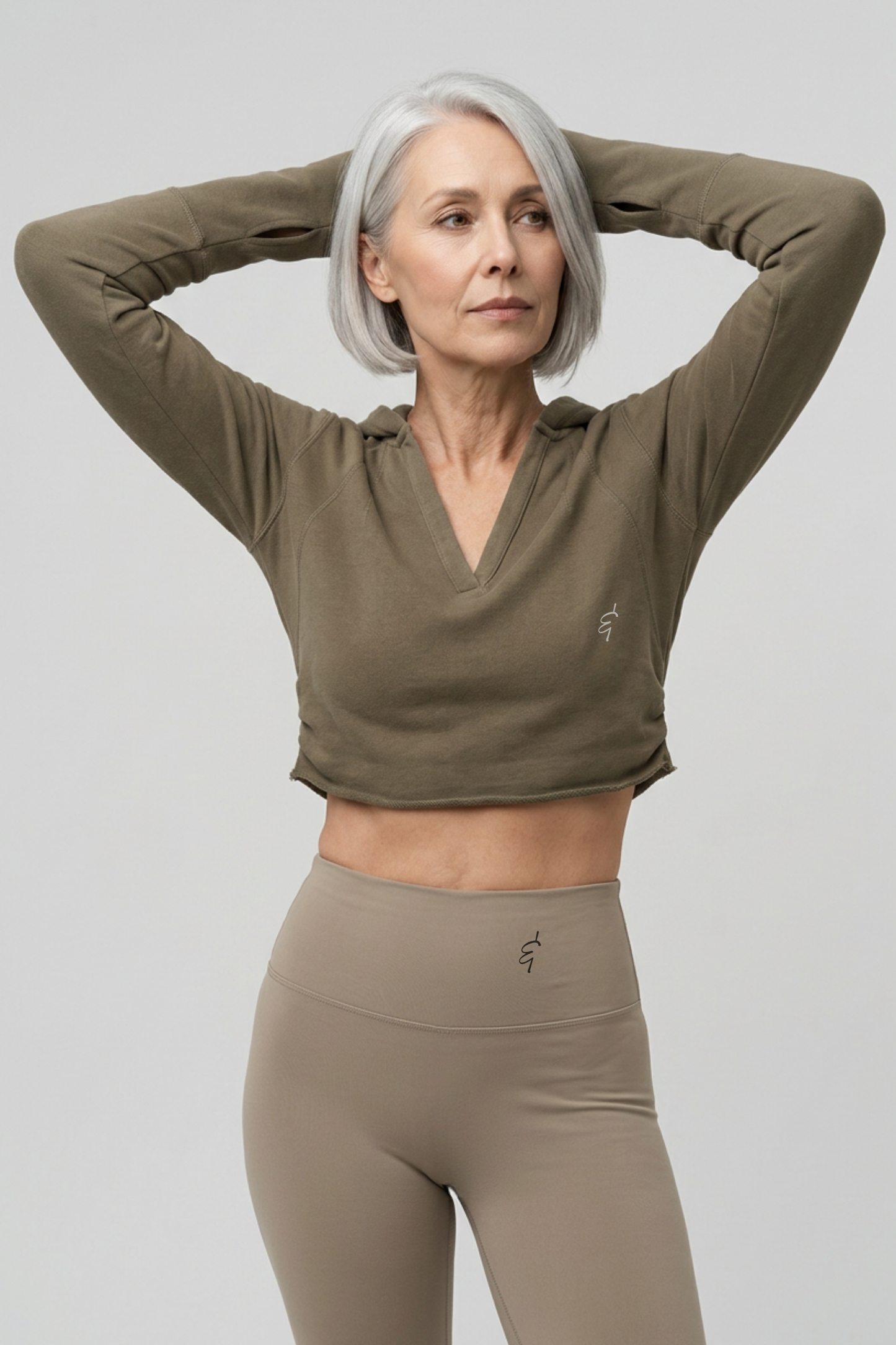 A woman with short gray hair poses in a cropped, long-sleeve athletic top and matching high-waisted leggings against a light gray studio background.