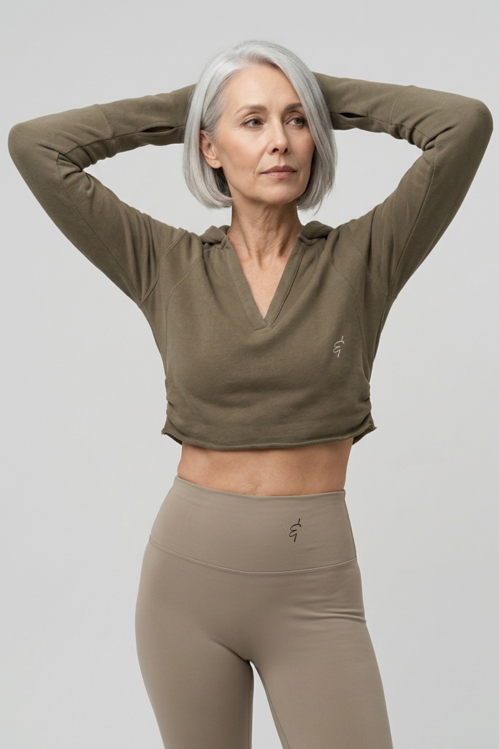 A woman with short gray hair poses in a cropped, long-sleeve athletic top and matching high-waisted leggings against a light gray studio background.