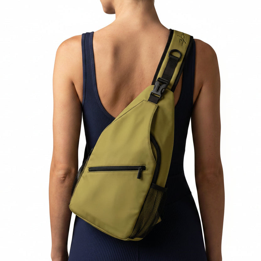A woman shown from behind wearing a navy blue athletic one-piece swimsuit and carrying a light olive green sling backpack across her shoulder, set against a solid warm gray studio background.