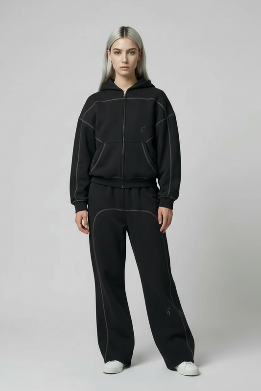 Woman wearing a black zip-up hoodie and matching joggers with dark gray contrast stitching along seams and pockets in fleece material