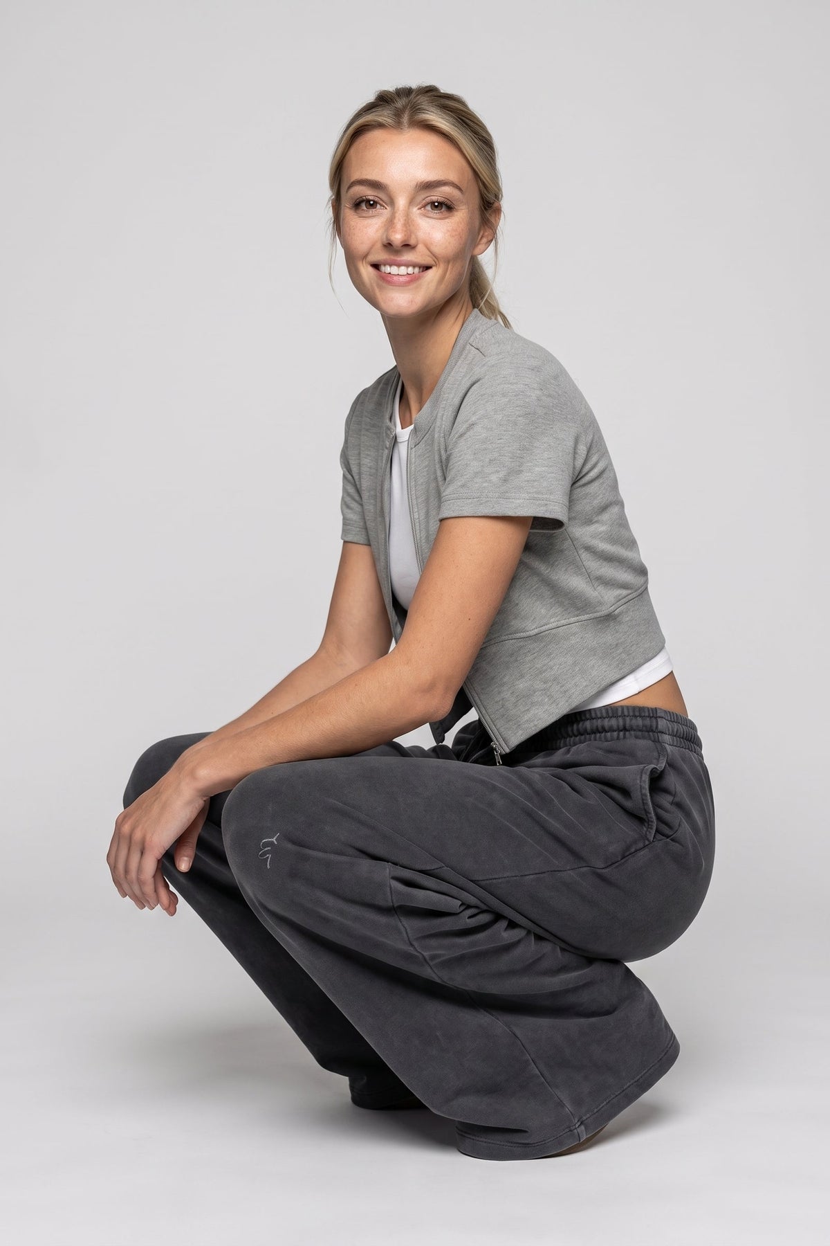 A woman crouching in a studio setting, wearing a light gray cropped short-sleeve top over a white undershirt and dark charcoal gray wide-leg pants.