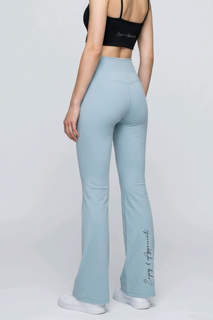 A woman stands in a studio wearing light blue flared leggings and a black sports bra, photographed from the back against a light gray background.