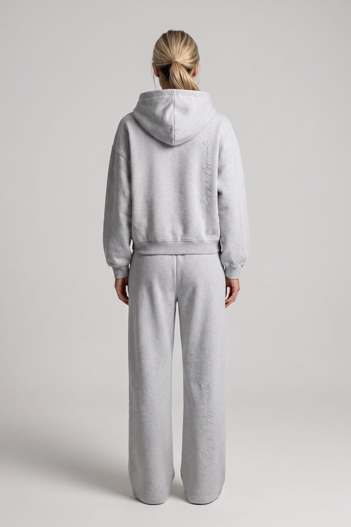 A woman viewed from behind wearing a light gray heathered matching loungewear set consisting of a cropped pullover hoodie and wide-leg sweatpants, shot against a neutral light gray studio background.