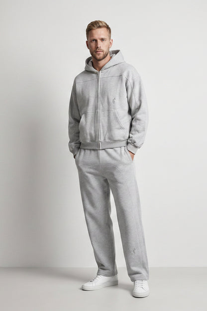 A man modeling a light gray zip-up hoodie and matching sweatpants set, standing against a neutral light gray studio background.