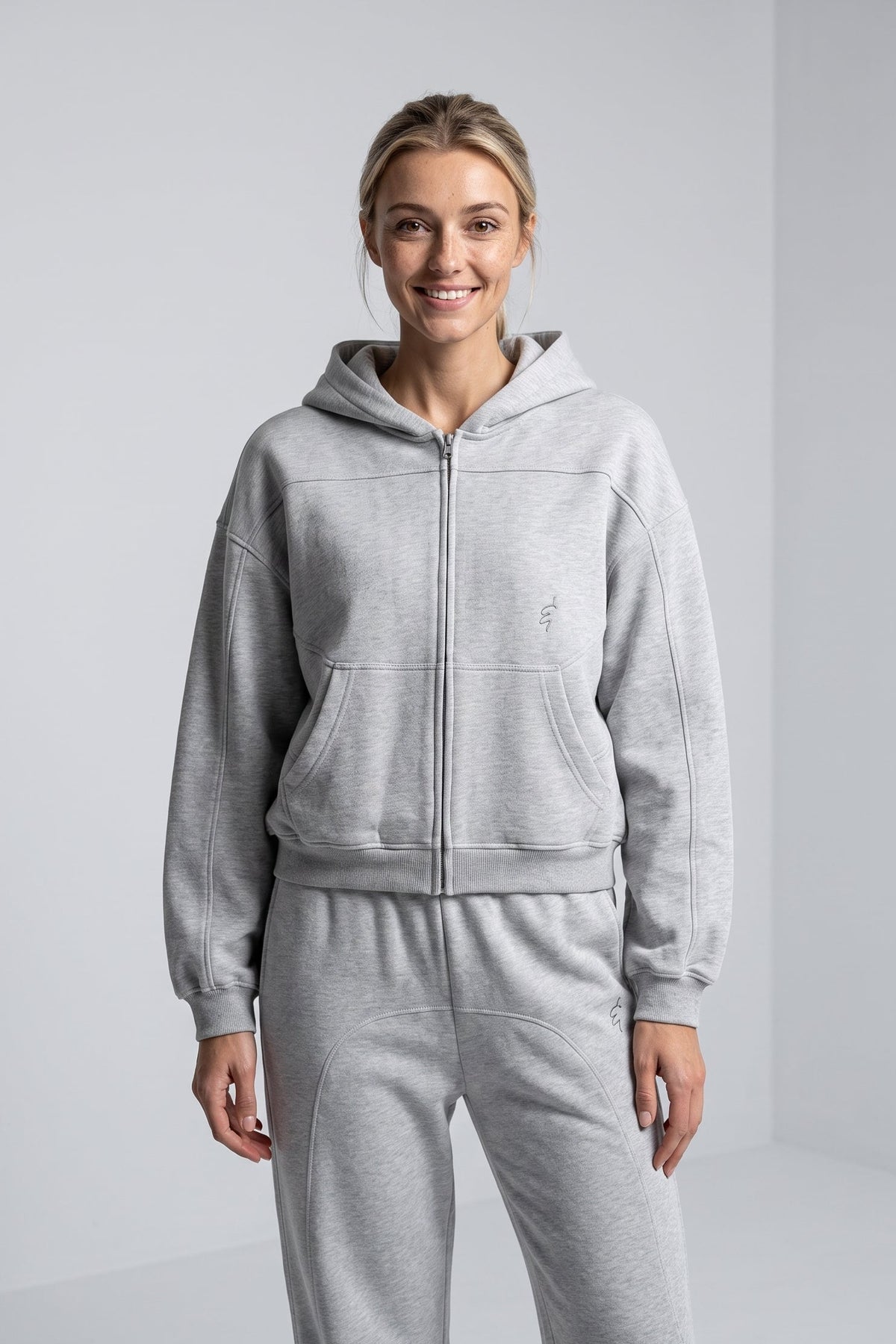 A woman modeling a light gray heathered zip-up hoodie and matching sweatpants set against a clean, light gray studio background.