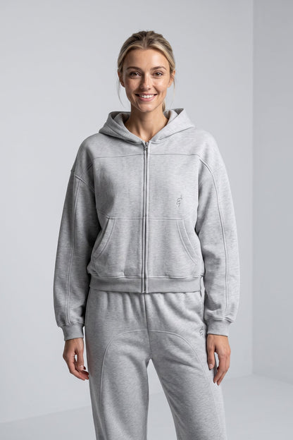 A woman modeling a light gray heathered zip-up hoodie and matching sweatpants set against a clean, light gray studio background.