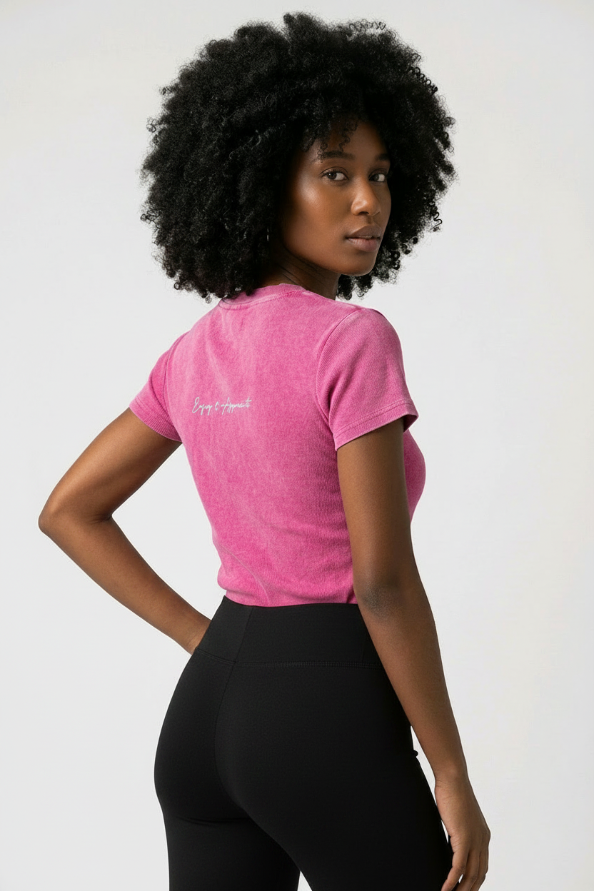 A woman with a large black afro is shown from the back wearing a bright pink heathered t-shirt with a small white script logo on the left shoulder, paired with dark blue jeans, against a plain light gray background.