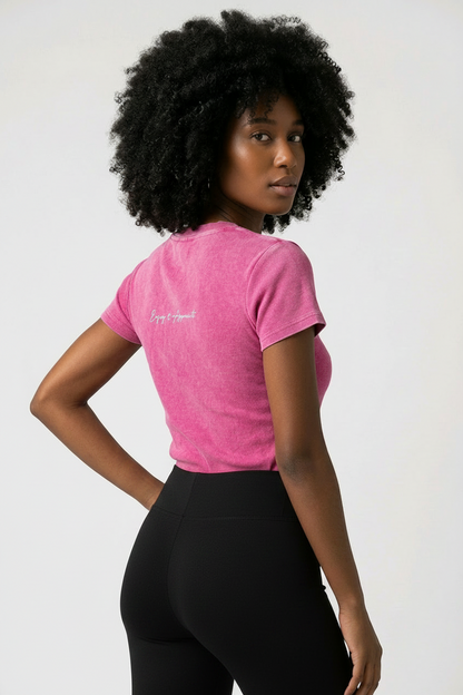 A woman with a large black afro is shown from the back wearing a bright pink heathered t-shirt with a small white script logo on the left shoulder, paired with dark blue jeans, against a plain light gray background.