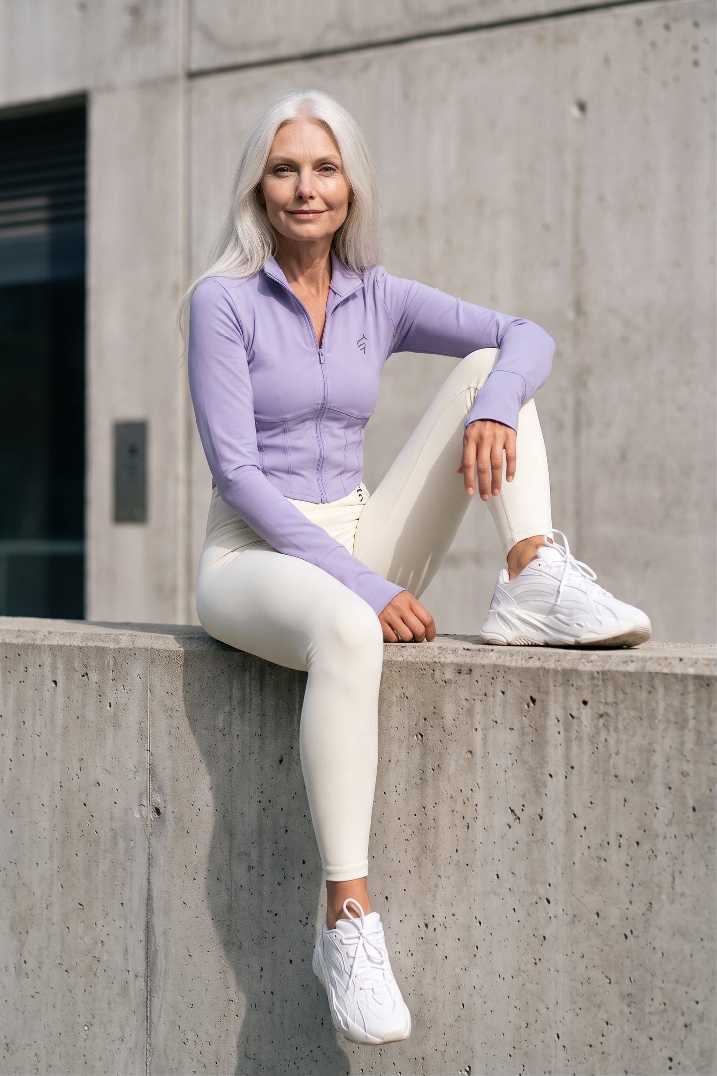 A woman with long white hair sits on a concrete ledge wearing a light purple long-sleeve athletic zip-up top, white leggings, and white sneakers against a concrete wall background.