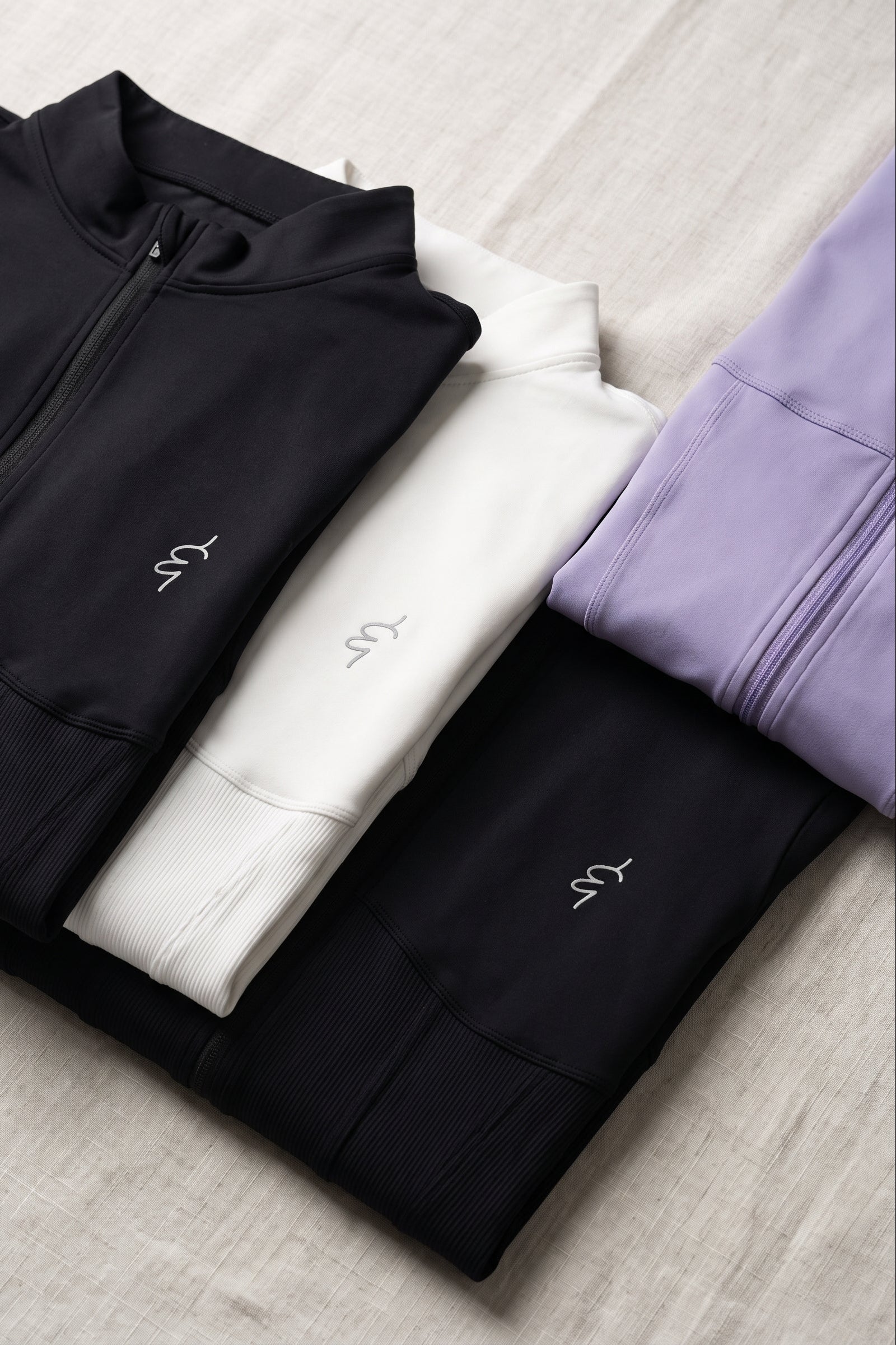 Three folded athletic zip-up jackets in black, white, and lavender purple, arranged diagonally on a light beige textured fabric background. Each jacket features a small white embroidered logo on the chest.