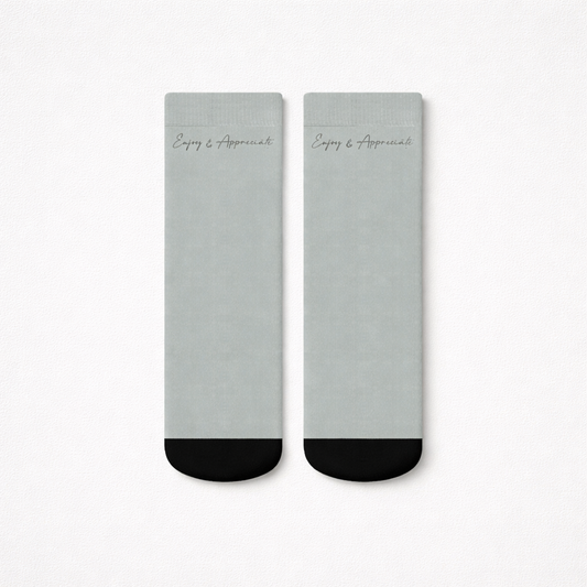 A pair of minimalist gray ankle socks with black toe caps and silver enjoy and appreciate script near the top
