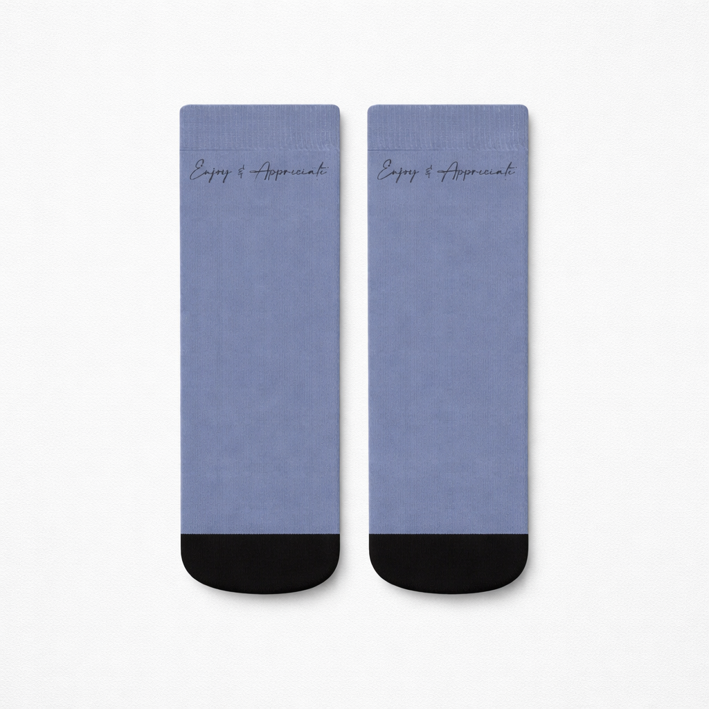 A pair of light blue ankle socks with black toe caps and enjoy and appreciate script near the top