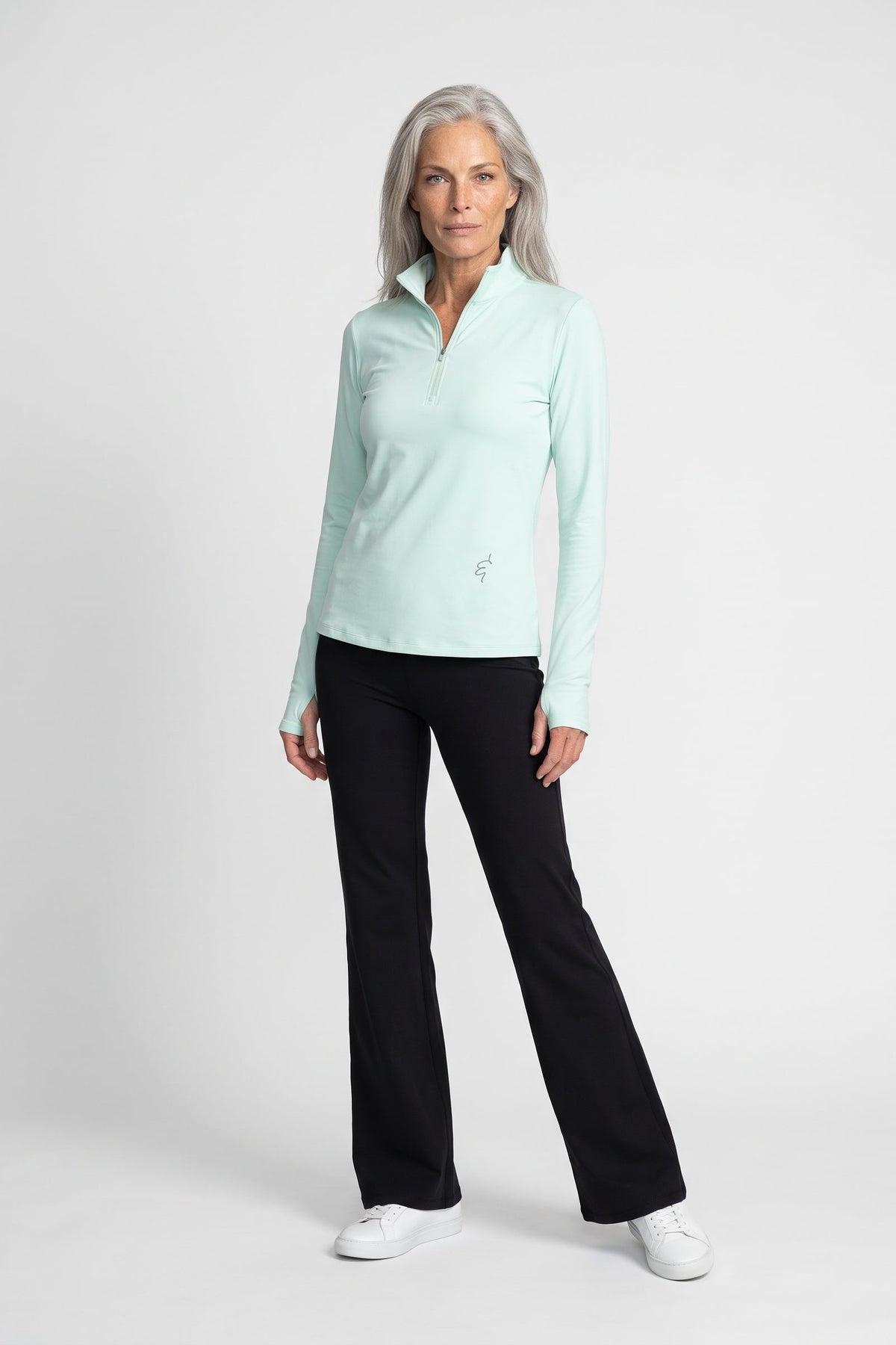 A woman modeling a light mint green long-sleeve quarter-zip pullover with black pants and white sneakers, standing against a light gray studio background.