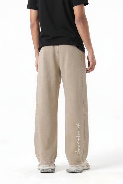 A model shown from the back wearing beige wide-leg sweatpants with a white script logo on the lower left leg, photographed against a clean white background.