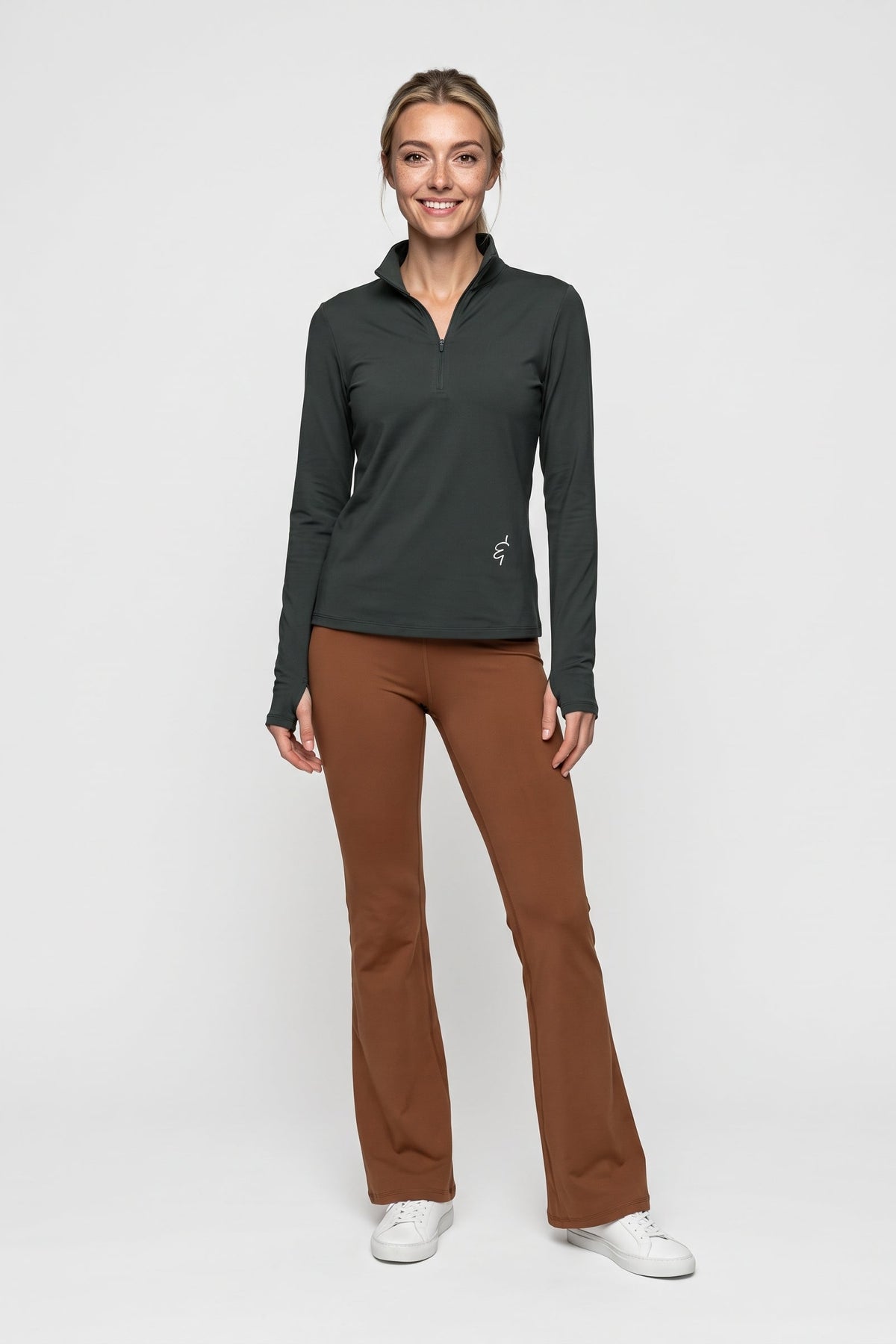 A woman modeling a dark charcoal gray long-sleeve quarter-zip athletic top paired with brown pants and white sneakers, standing against a light gray studio background.