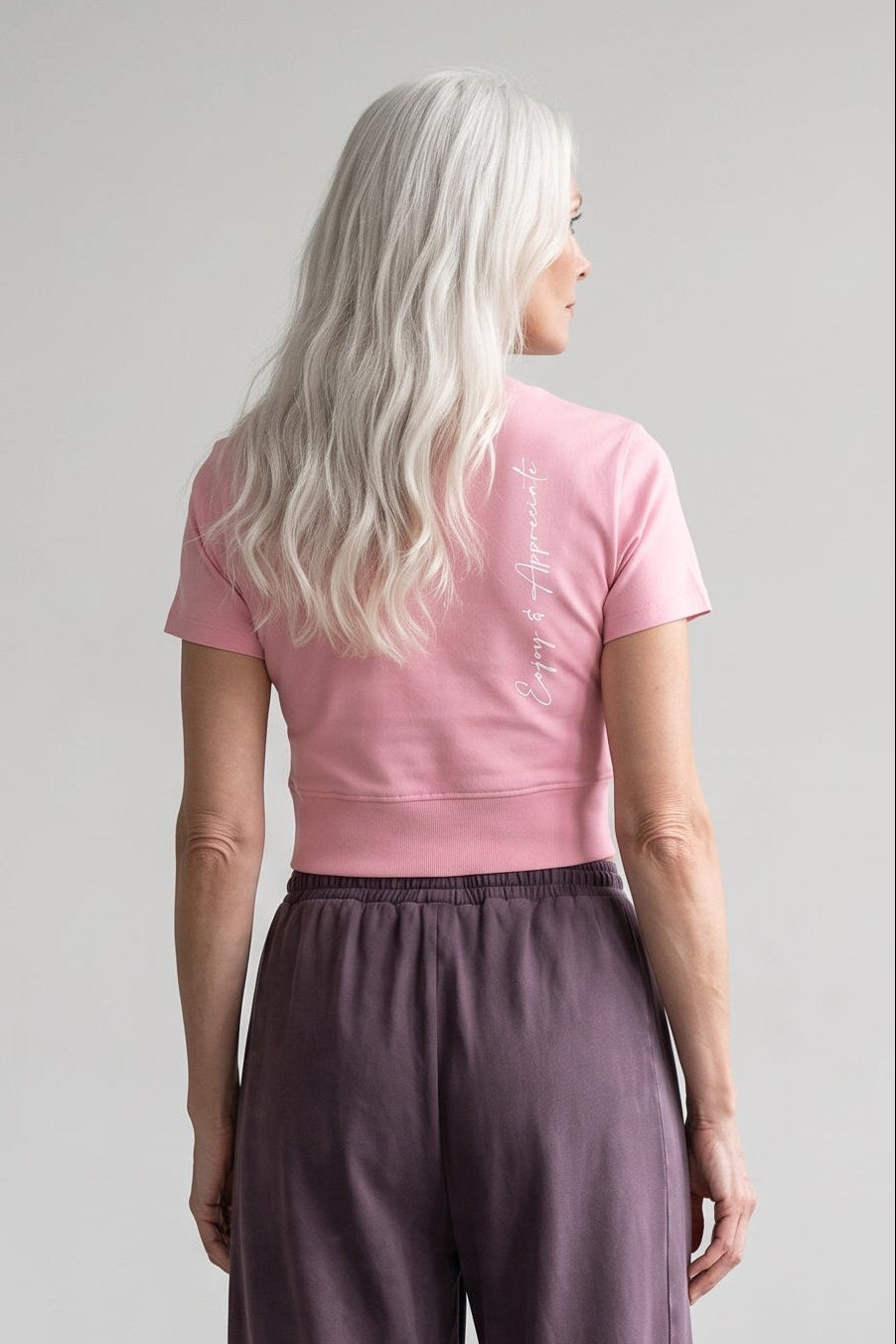 A woman with long platinum blonde hair is shown from behind wearing wide-leg mauve-purple trousers and a pink t-shirt, standing in a minimalist studio setting.