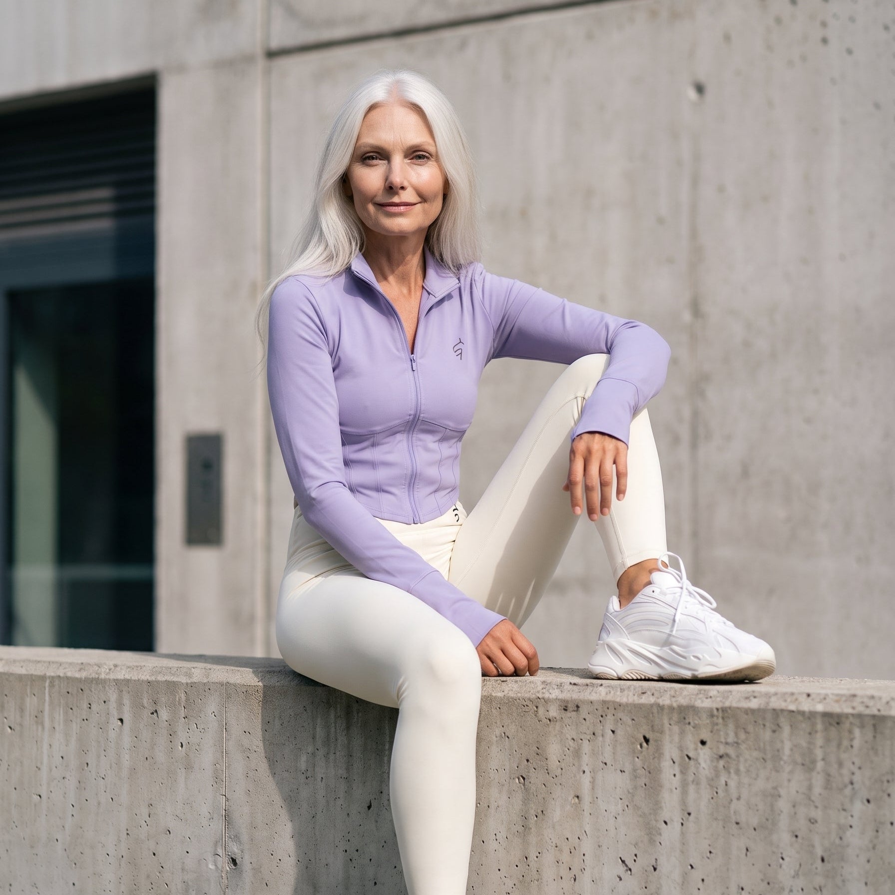 A woman with long white hair sits on a concrete ledge wearing a light purple long-sleeve athletic zip-up top, white leggings, and white sneakers against a concrete wall background.