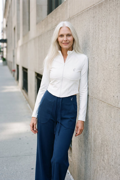 A woman with long white hair stands against a concrete building wall, wearing a white long-sleeve zip-front top and dark navy wide-leg pants.