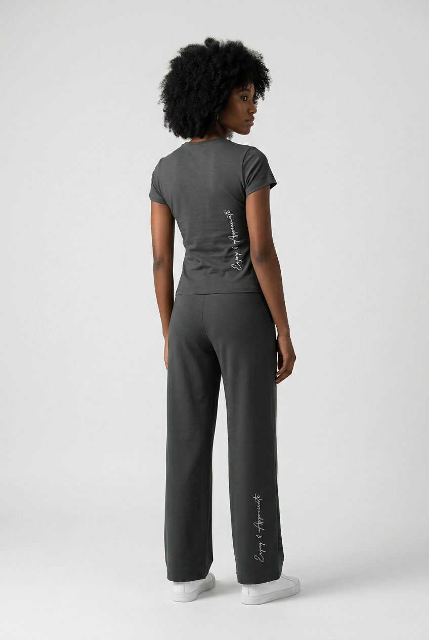 A model shown from the back wearing a dark gray short-sleeve t-shirt and matching wide-leg sweatpants with white script text down the back and left leg, photographed against a light gray studio background.