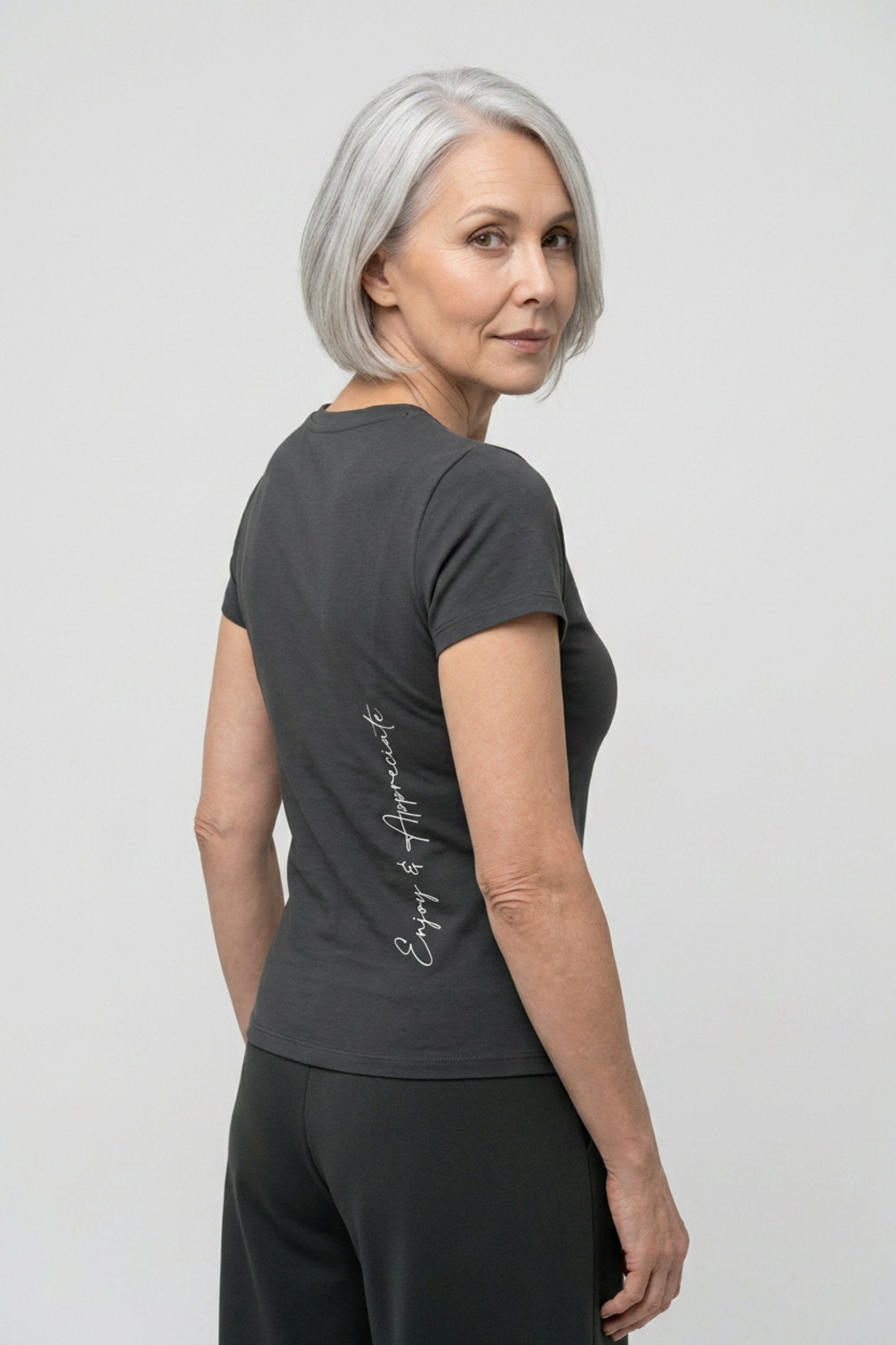 An older woman with short gray hair is shown from the back wearing a dark charcoal gray t-shirt with 'Enjoy & Appropriate' printed in white script down the back, paired with dark gray pants, against a light gray studio background.