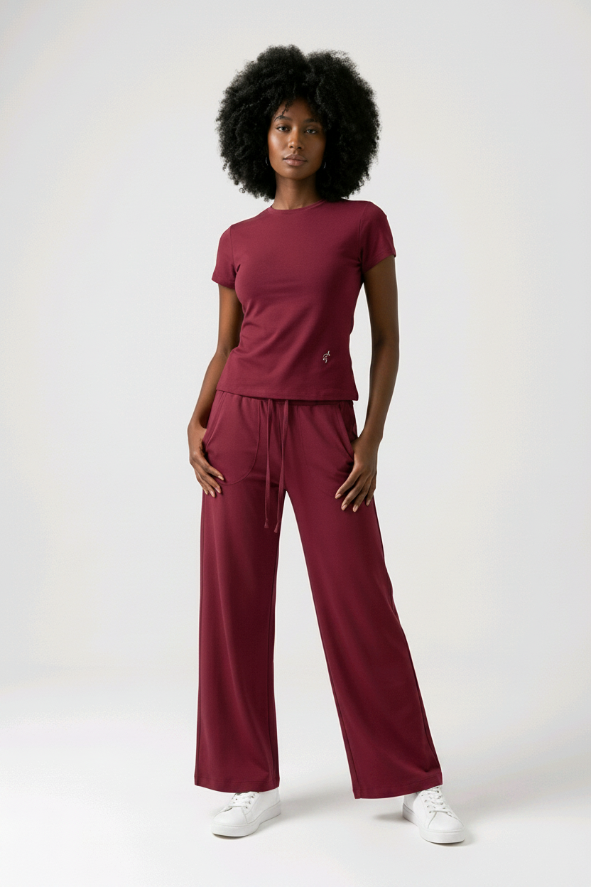A woman modeling a matching burgundy scrub set consisting of a short-sleeve top and wide-leg pants, paired with white trainers, against a plain light gray background.